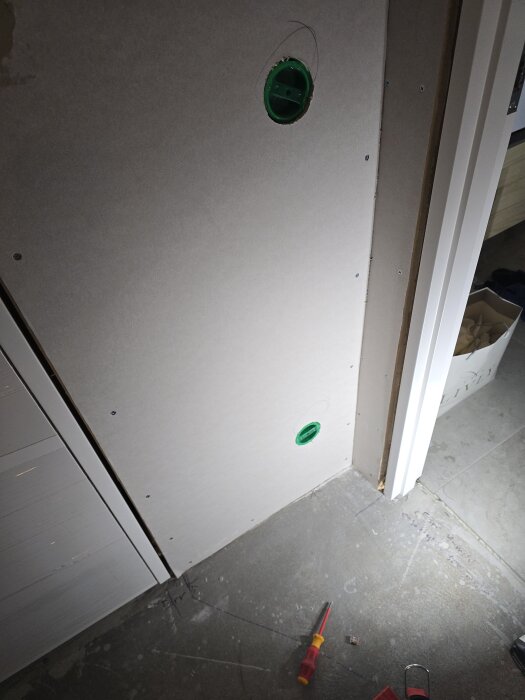 Vägg i konstruktion, två gröna eluttag synliga i gipsväggen, skruvmejsel och kabel på betonggolv, bredvid en dörröppning.