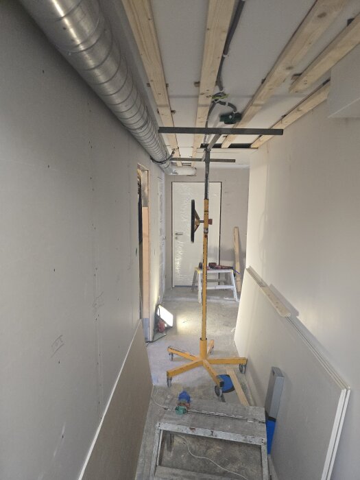 Renoveringskorridor med pågående arbete, träreglar i taket, synlig ventilation, bygglampor och stege längs väggen. Förberedelse för gipsning.