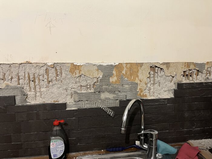 Porös vägg bakom kökskran med avlägsnat kakel, blottad armering av vass, och kvarvarande puts i en pågående köksrenovering.