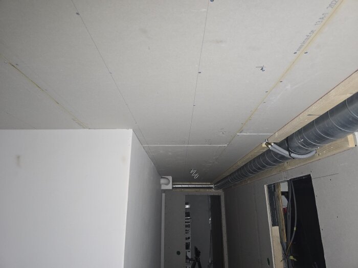 Gipstak och väggar i en källarkorridor under renovering, med synliga ventilationsrör och kablar innan slutlig ytbearbetning.