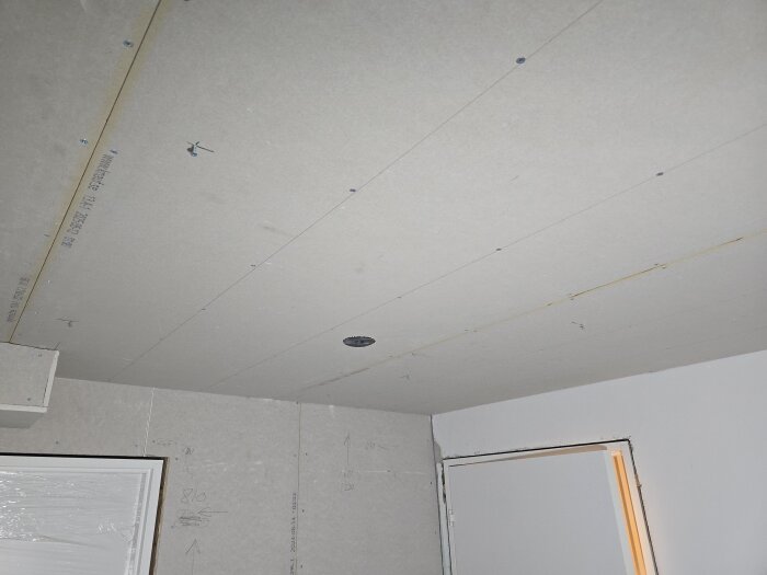 Nymonterat gipstak med infällda takdosor i en källarkorridor under pågående renovering, vägg med dörr syns till höger.