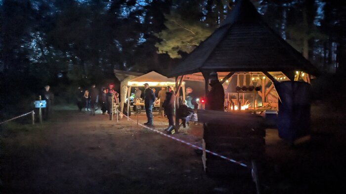 Utomhusevenemang på kvällen med människor samlade runt ett upplyst tält och en eldplats under ett tak.