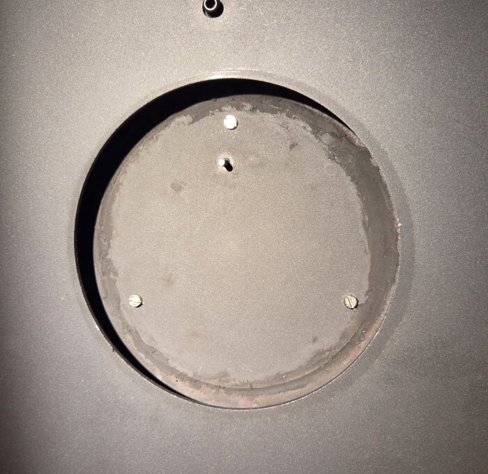 Rund öppning i metall med tre synliga skruvar och mörkare kitt runt kanterna, omgiven av en ljusare bakgrundsyta.