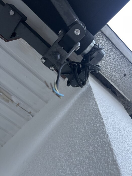 Närbild av en markisarm med exponerade elsladdar, monterad på en vit strukturerad vägg under ett tak med vit yta och synliga skruvar.