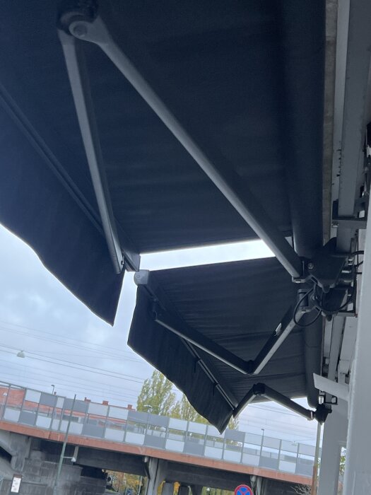 Svart markis med metallstöd monterad vid en byggnad, med väg och bro i bakgrunden under en molnig himmel.