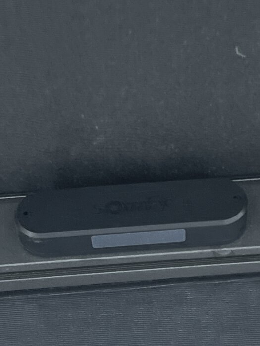 Svart enhet monterad på en fönsterkarm, möjligtvis en sensor eller kontrollmodul med text och en liten skärm eller LED-indikator på framsidan.