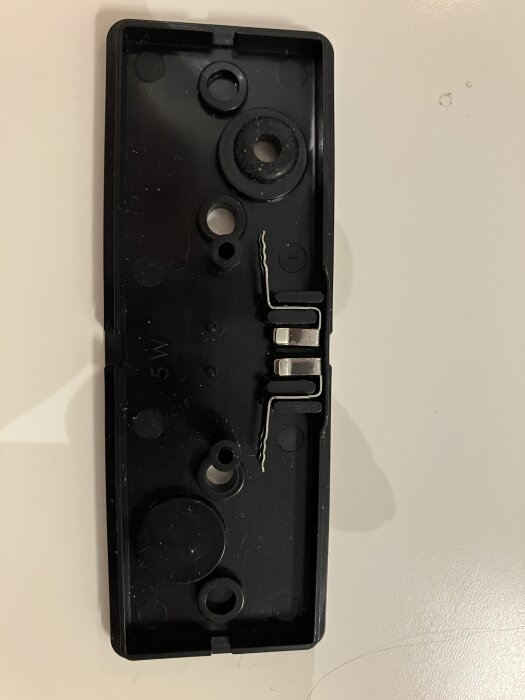 Svart plastdel med flera runda fästhål och metallkontakter, möjligen från en positionslykta.