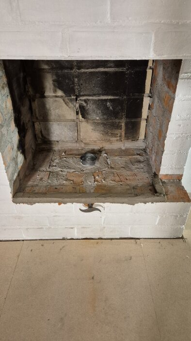 Öppen eldstad med svartnade tegelstenar och ett ventilationsrör i botten, omgiven av vitmålad tegelvägg.