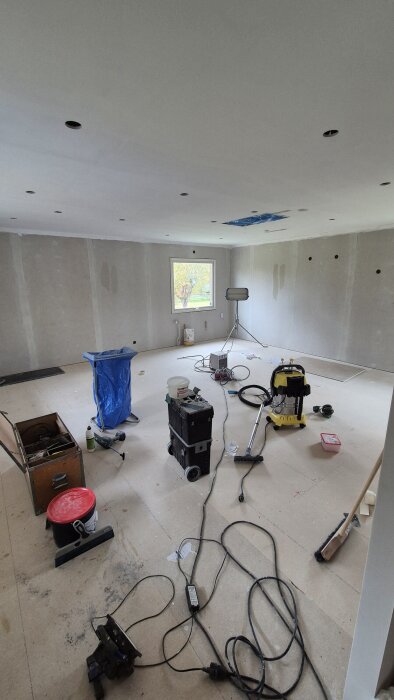 Ett rum under renovering med målat tak, verktyg, dammsugare och kablar på golvet. En lampa står vid fönstret.