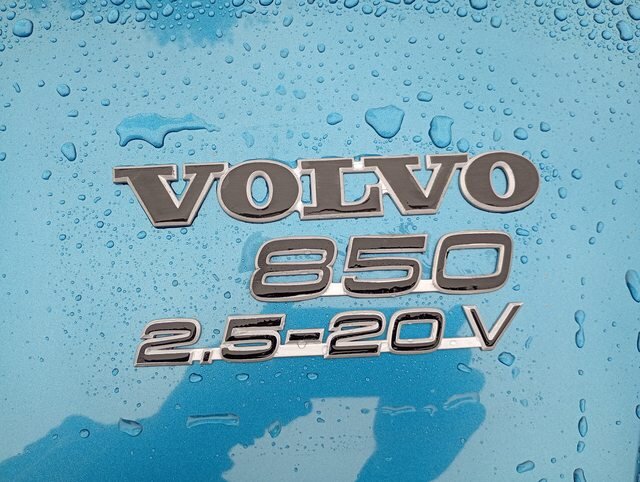 Volvo 850-emblem med silverkanter och svart mitt på en blöt blå bilhuv.