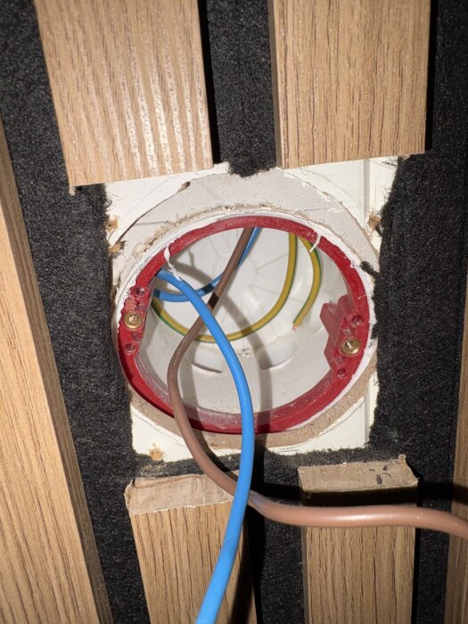 Väggmonterad apparatdosa med synliga elkablar, omgivet av träpanel och svart isolering, för diskussion om förhöjningsringar och montering.