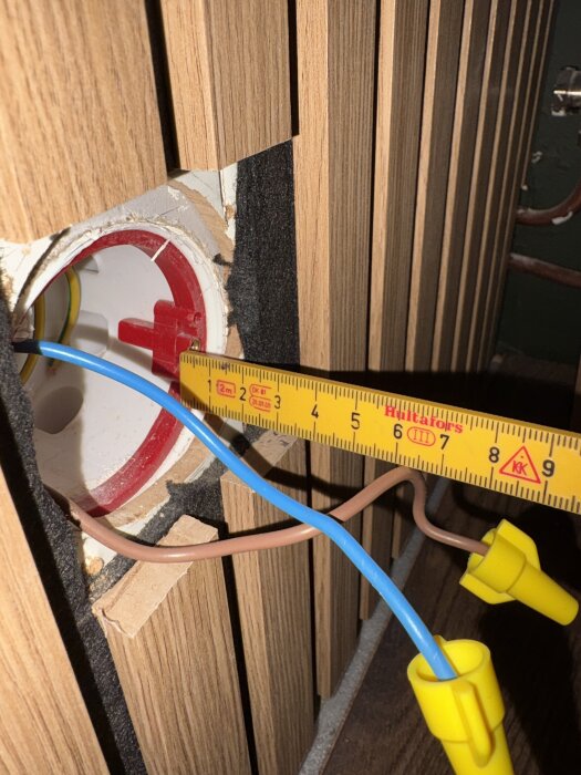 Elektrisk apparatdosa med blå och brun ledning samt hoprullat måttband, omgiven av träpanel, visar 30 mm djup.
