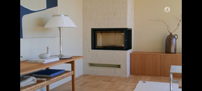 Rektangulär kakelspis i rum med trägolv, lampa på bord bredvid.