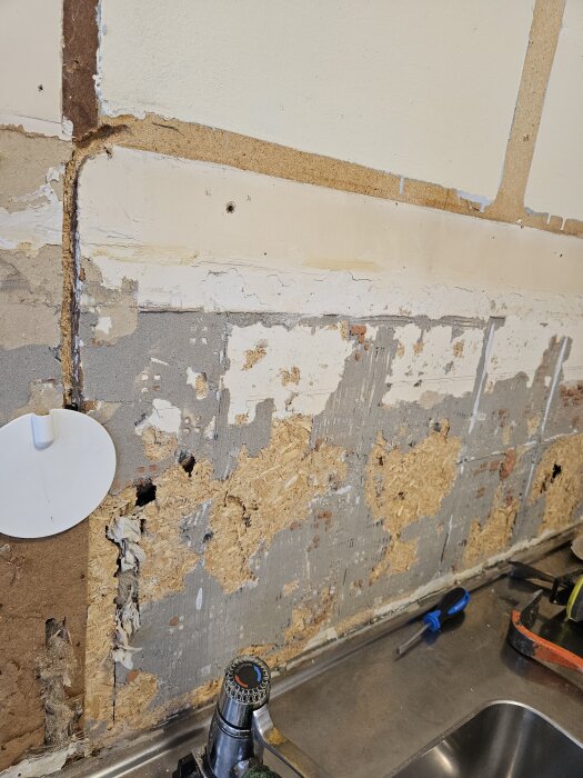 Ojämn och skadad köksvägg efter rivning, med synlig isolering och fuktspärr; verktyg synliga på bänken.