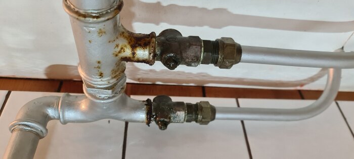 Rörsystem med två ventiler, några delar rostiga. Sprejat med WD40, syns på fuktigheten i bilden. Golvplattor och vägg synliga i bakgrunden.