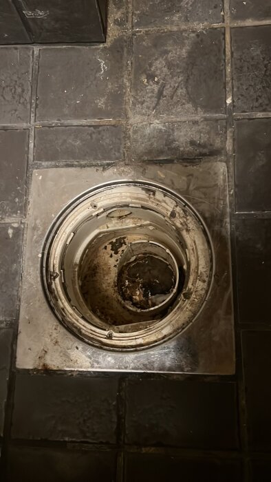 Används avlopp i tvättrum, utan vattenlås, med trasig insats. Brun smuts synlig runt öppning. Kakelgolv synligt runtom avloppet.