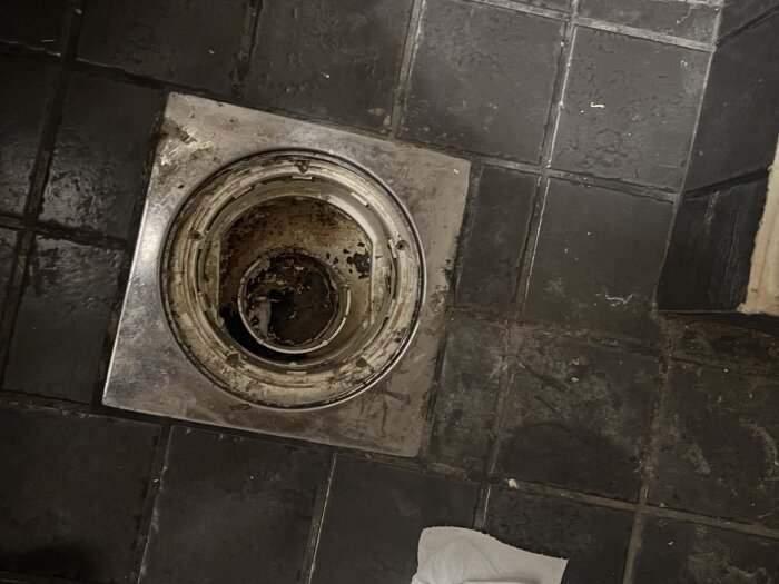 Trasigt golvbrunnsutlopp i tvättrum utan vattenlås, omgiven av svarta kakelplattor, delvis trasig insats synlig.