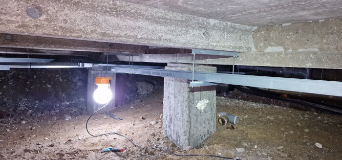 Krypgrund under hus med belysning, metallbalkar och verktyg på marken, för PEX-installation och rörsystembyte.