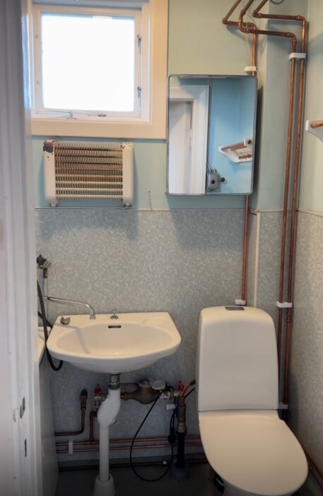 Gammalt badrum med väggfast handfat, toalettstol, spegel med skåp, väggmonterad radiator och synliga kopparrör, i ett 30-talshus.