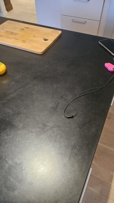 Köksbänk med skärbräda, en apelsin, en svart kabel samt en rosa klämma, med ljusa köksskåp i bakgrunden.