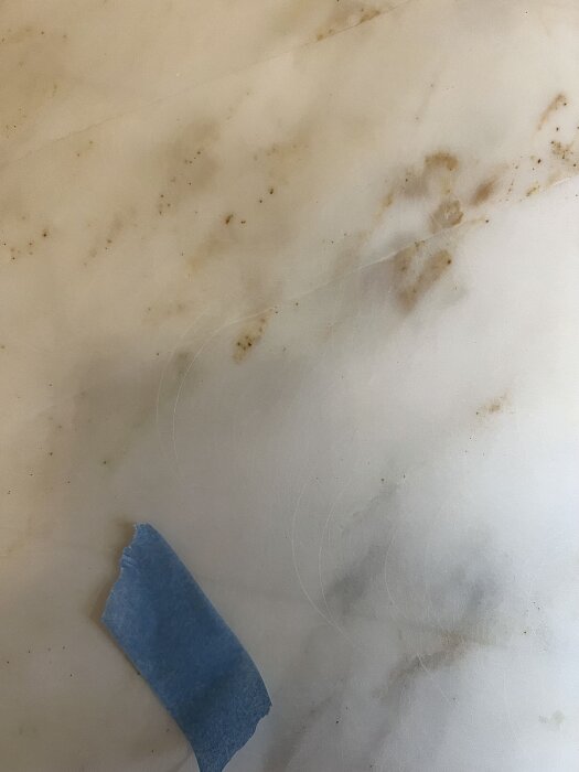 Marmorplatta med synliga repor och virvlar samt smuts i djupare ådring. Blå tejpbit finns på ytan.