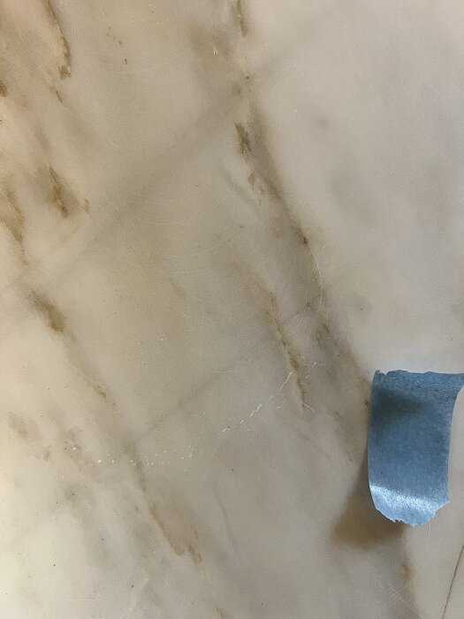 Marmor med repor och virvlar samt en bit blå tejp. Synlig ådring och tecken på felaktig slipning som orsakat ojämn yta.