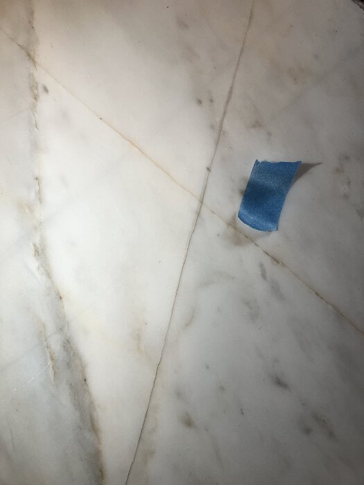 Marmorgolv med repor och smuts i ådring, blå tejpbit markerar ett område.