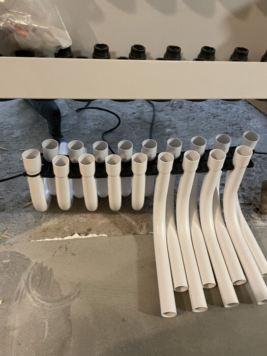 Vita böjar för golvvärmeslingor placerade under ett fördelarskåp på ett betonggolv, redo för installation och fastsättning i träreglar.