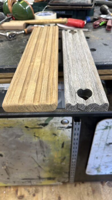 Två träplankor på en arbetsbänk, en behandlad med varm färg och en gråblekt, omgivna av verktyg och målartejp.