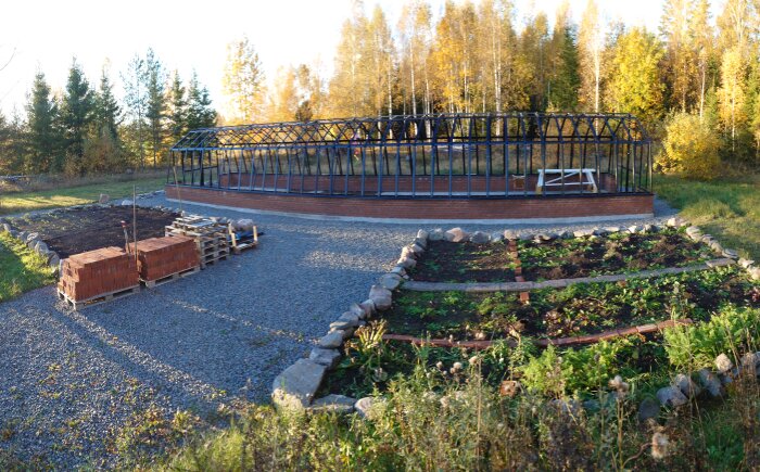 Växthus under konstruktion med metallram och murverk, omgiven av höstfärgade träd och odling, med staplade tegel på pallar i förgrunden.