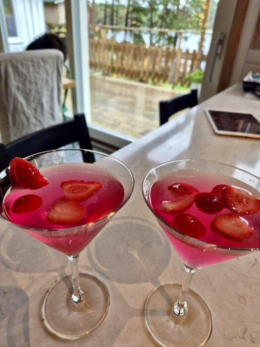Två glas med rosa raspberry sour-cocktail, dekorerade med skivade jordgubbar, står på ett bord med utsikt över en regnig trädgård.