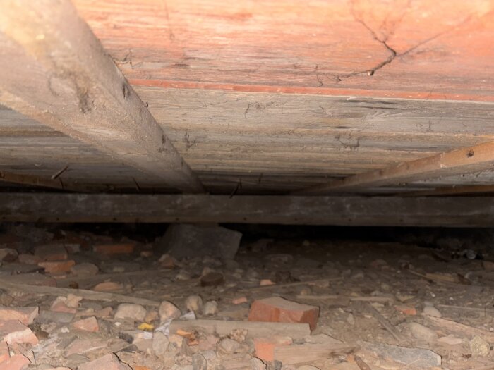 Krypgrund med tegelrester och synliga träbalkar ovan, sedd genom ventilationshål under plintgrunden.