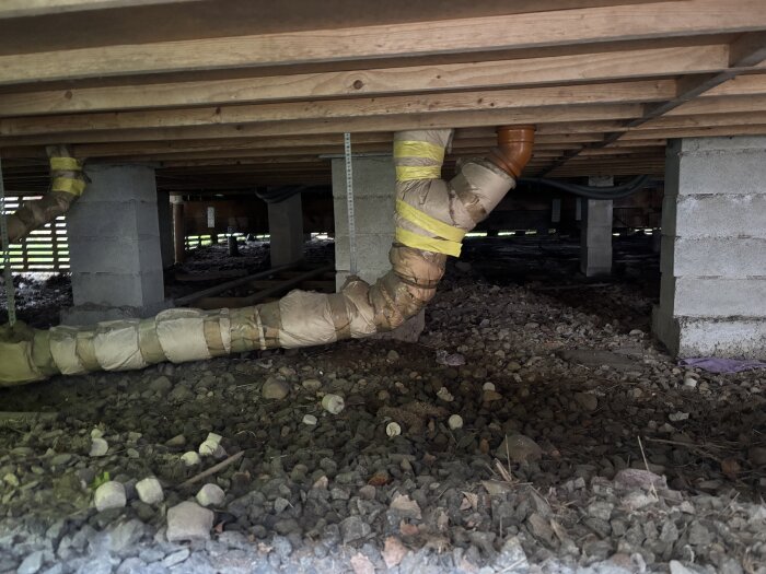 Öppen krypgrund med ventilationsrör och plintar. Markyta täckt av sten och grus.