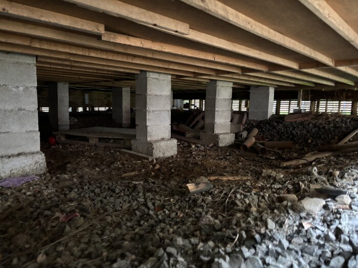 Öppet utrymme under plintgrund med synliga pelare, rör och stenar i en krypgrund med ventilationshål.