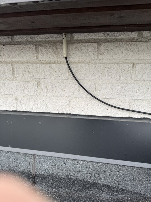 Kabel som går längs garageväggen från marken, fastsatt i en vit kopplingsbox under taket, stenbelagd fasad i bakgrunden.