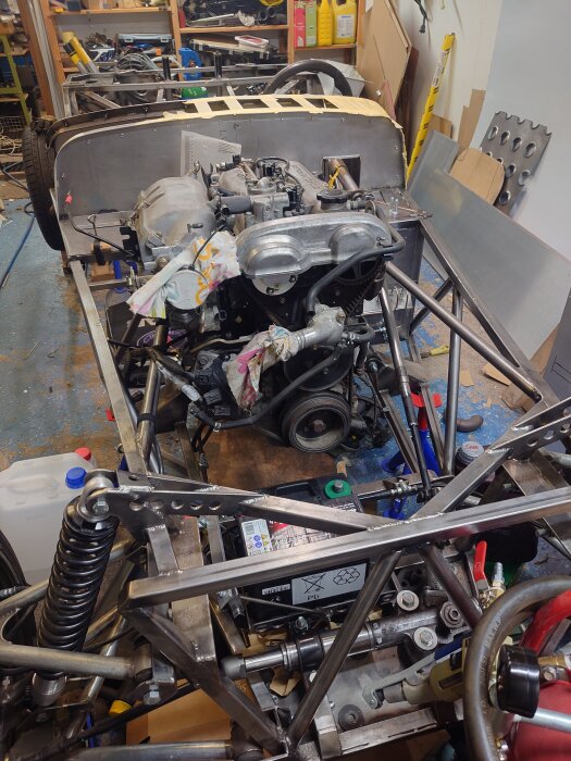 Motor och chassi av en bil under konstruktion, troligen en Miata NB med 1,8 liters radfyra, omgiven av verktyg och byggmaterial i ett garage.