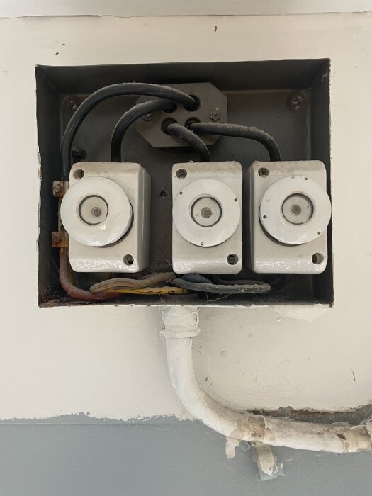 Öppen elskåp med tre gamla keramiska säkringar och ledningar i ett metallhölje, monterat på en vägg.