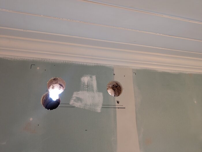 Två borrade hål i taknära vägg med spår av verktyg kring kanterna, belysta av infallande dagsljus.