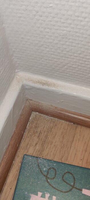 Vägg i hörn med träspån och damm på golv och list, närbild tagen vid borrningsarbete för hyllmontering.