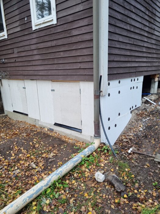 Husfasad med isolerade lecablock och cellplast, brun träpanel ovanför; diskuteras om att putsa eller använda fibercement. Marken täckt med löv.