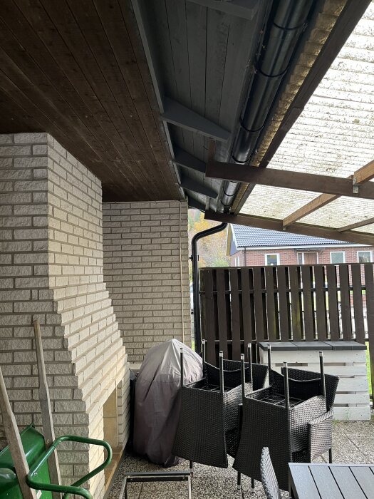 Altantak i trä och plast fäst vid tegelvägg. Rännor och pelare synliga, staplade stolar bredvid en täckt grill under taket.