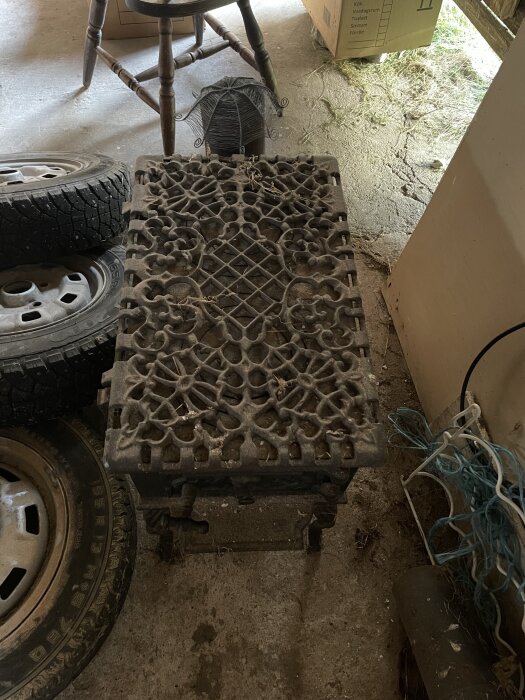 Dekorativ gjutjärnskamin med detaljerat mönster, omgiven av däck, stol och kartonger i ett garage eller verkstad.