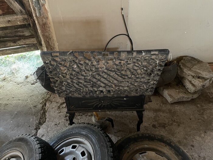 Gjutjärnskamin under renovering med dekorativ topp. Står i ett garage med gamla däck runtomkring.