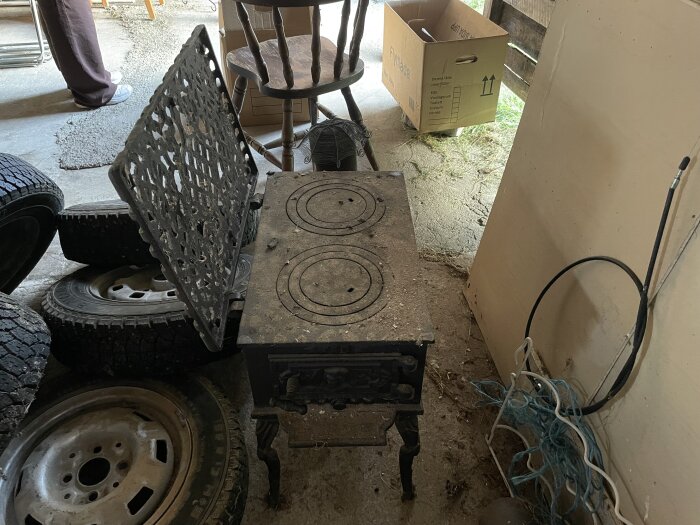 Gjutjärnskamin med utskuren lucka uppfälld, omgiven av gamla bildäck och en flyttlåda i en verkstad.