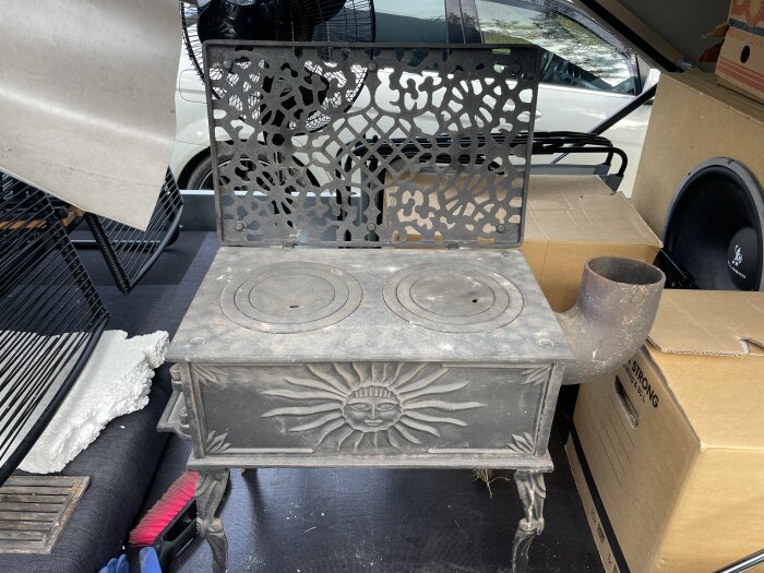 Gammal gjutjärnsspis med solmotiv och dekorativt hålmönster, redo för putsning, placerad i bilen bland kartonger och borste.