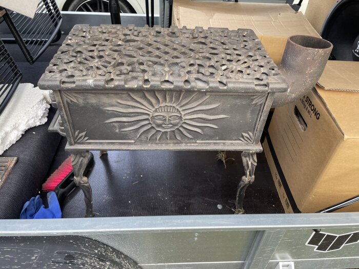 En rengjord gjutjärnsspis med utsmyckad soldekoration på sidan, redo för polering, står på ett underlag med kartonger i bakgrunden.