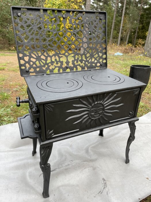 Svart kamin med solmotiv och ornerad lucka, stående utomhus på ett vitt underlag.