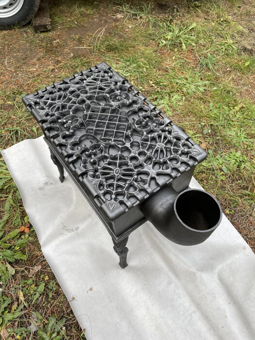 Svart, dekorativ liten kamin med mönstrat lock, placerad på vit duk utomhus på gräs.
