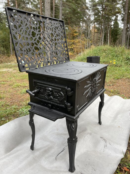 Liten svart kamin med dekorerat lock och ben står på en plats utomhus.