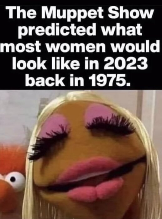 En Muppet-karaktär med blonda peruk och sminkade läppar och ögonfransar, med ett annat Muppet-ansikte suddigt i bakgrunden.